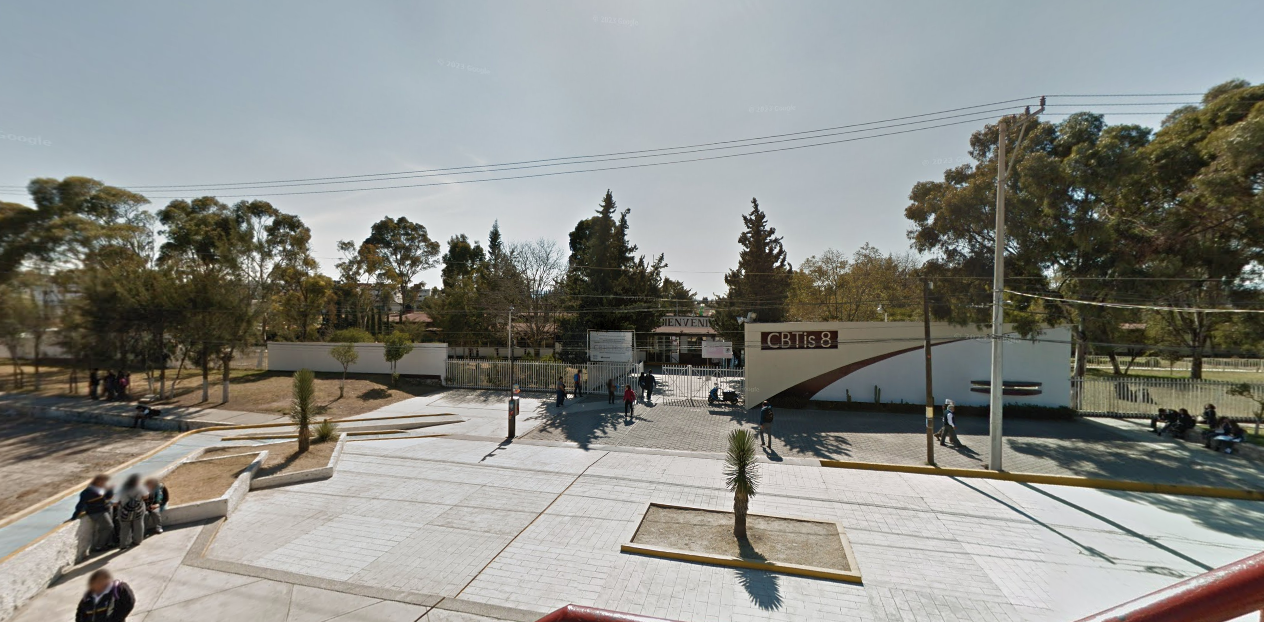 Fotografía en la que se muestra una explanada amplia. Al terminar la explanada está la entrada del plantel, conformada por una reja. A lado derecho de la reja hay un muro amarillo. Sobre el muro hay una placa de color rojo que dice en letras grandes Cebetis 8